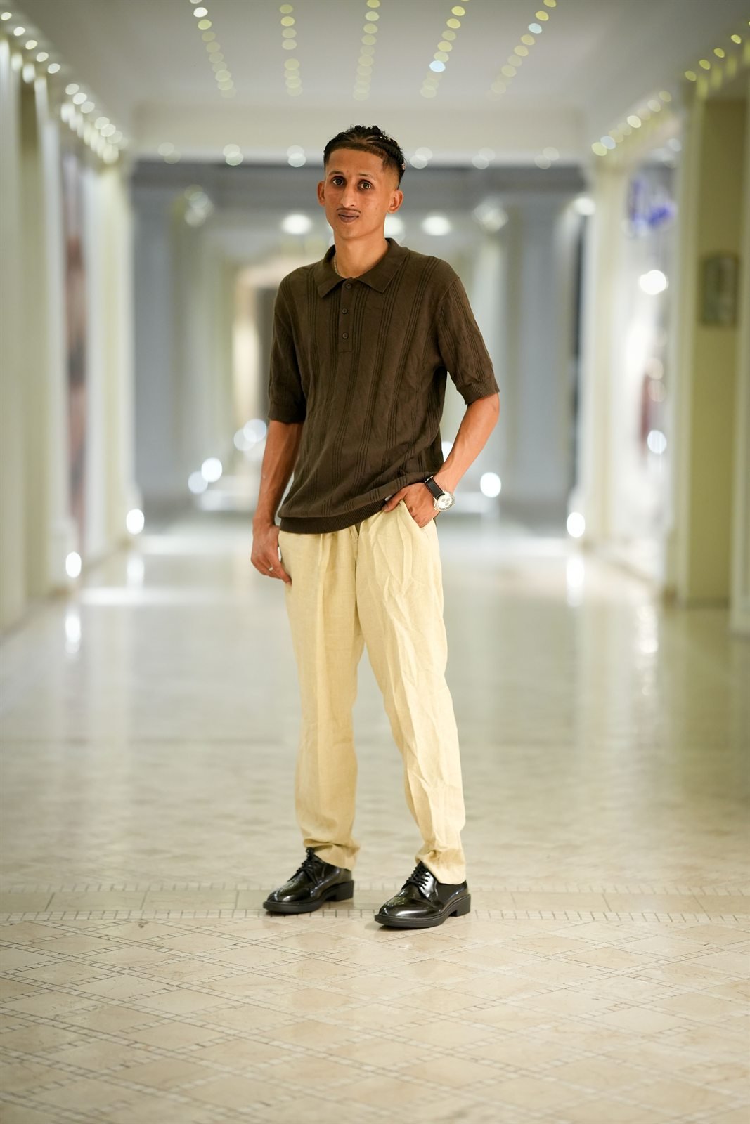 Fida Nadeem a figura intera in un corridoio illuminato, polo marrone e pantaloni chiari